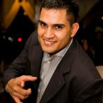 Portrait of a confident, smiling businessman in a formal suit pointing directly at the camera indoors.